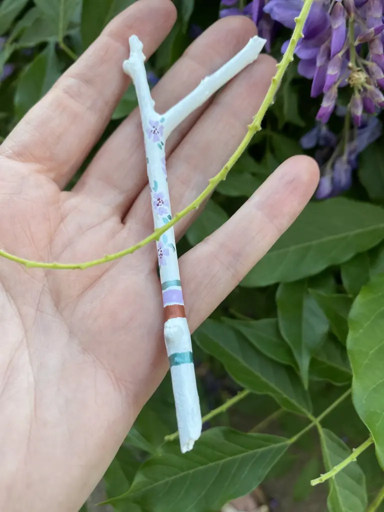 petit bâton de parole de 11 cm avec une fourche peint en blanc avec 4 bandes de couleurs vert sauge métallisé, cuivre et mauve puis 8 petites fleurs délicates peintes sur la partie supérieure.